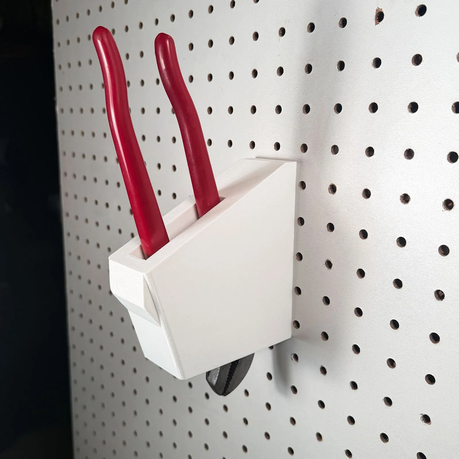 A white 3D printed holder on white pegboard with a pair of red-handled channel lock pliers standing in it, handles up and leaning away from the board, and the steel jaws showing below the holder