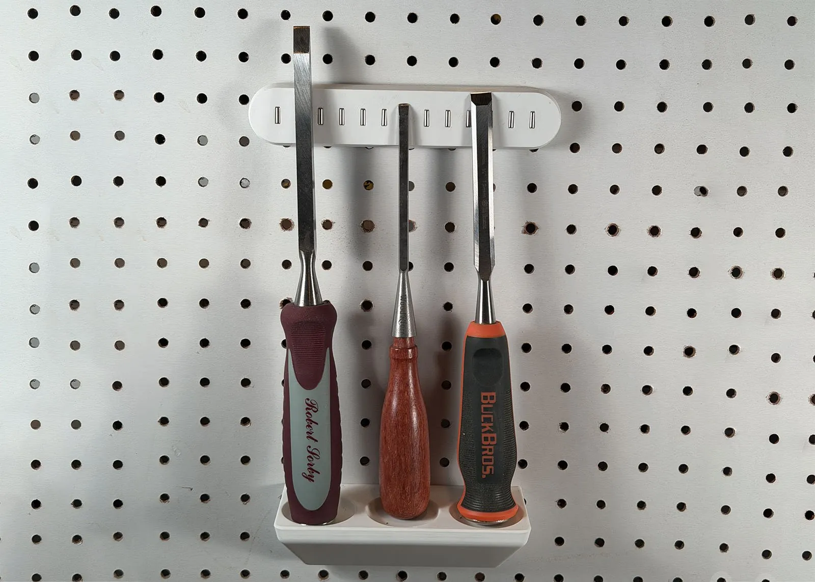 Front view of the rack and the magnet bar on pegboard, with the bar centered over the rack near the tops of the three chisel blades