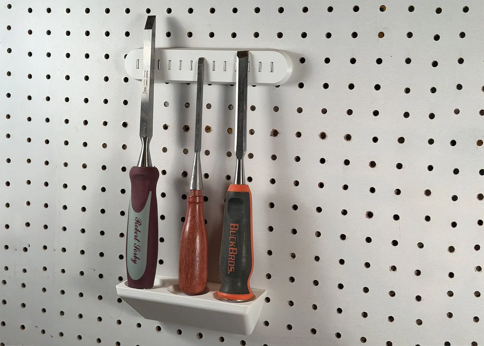 A white 3D printed rack on white pegboard holding three chisels of three different makes, with their handle ends in countersinks and their blades leaning back on a white magnetic bar higher up the board
