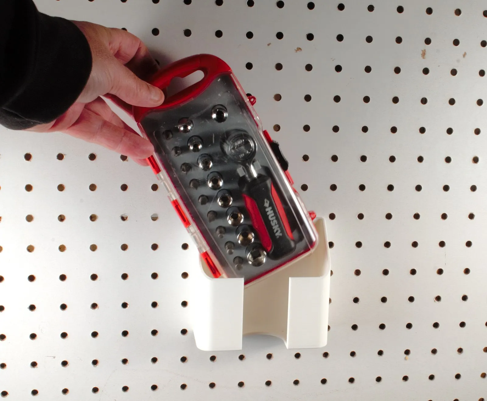 A hand lifting the driver set case out of the pegboard holder while the holder stays screwed to the board