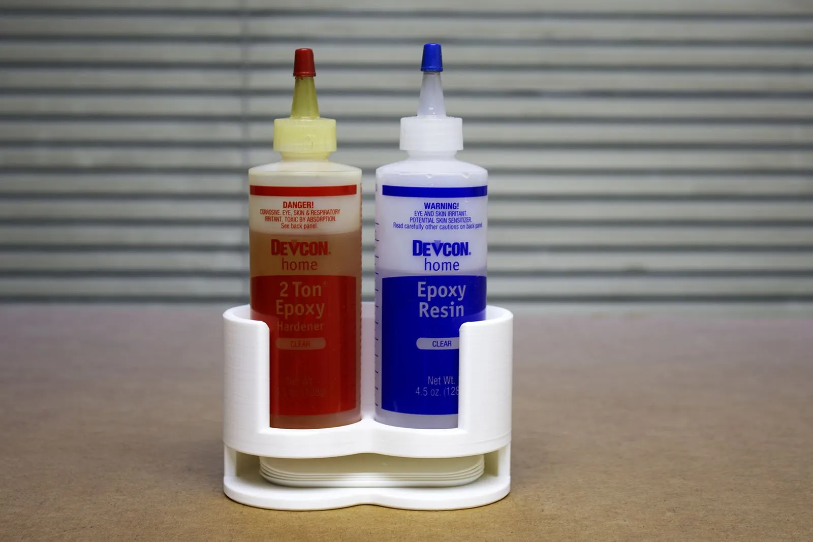 The rack standing on a workbench holding two Devcon epoxy bottles, the 2 Ton Epoxy Hardener in red and the Epoxy Resin in blue, with mixing trays stacked underneath