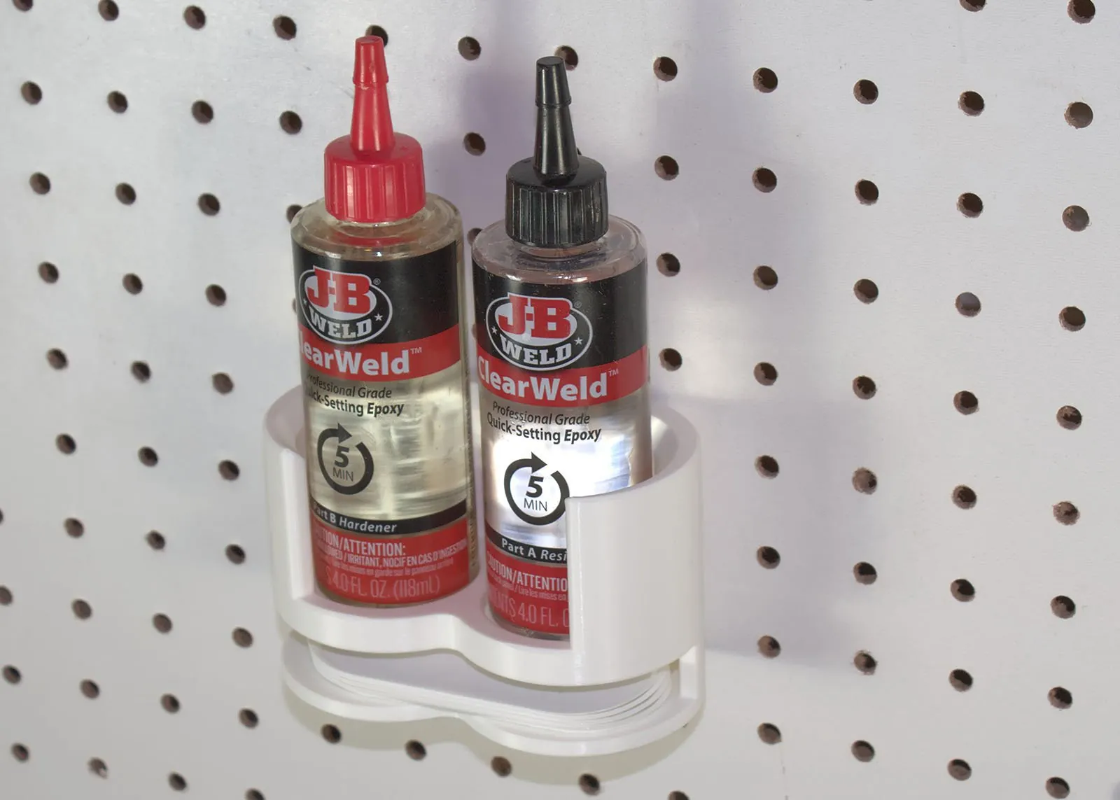 A white 3D printed rack on white pegboard holding two J-B Weld ClearWeld epoxy bottles, Part B with a red cap and Part A with a black cap, with a stack of mixing trays in a slot under the bottles