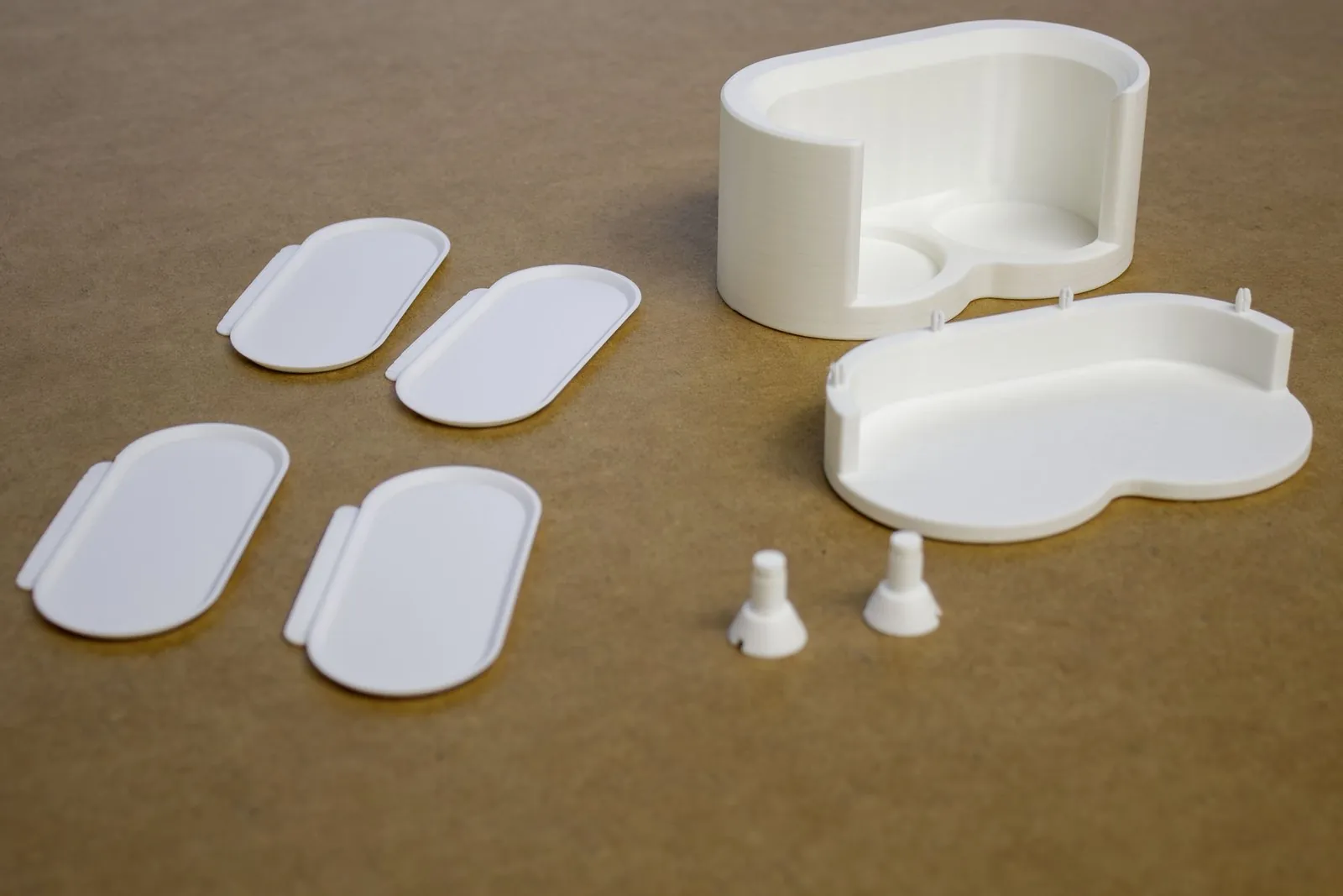 All the printed parts before assembly on a brown work surface: four oval mixing trays with tabs, the top piece with the two bottle pockets, the bottom piece with four small snap pins on its back wall, and two screws