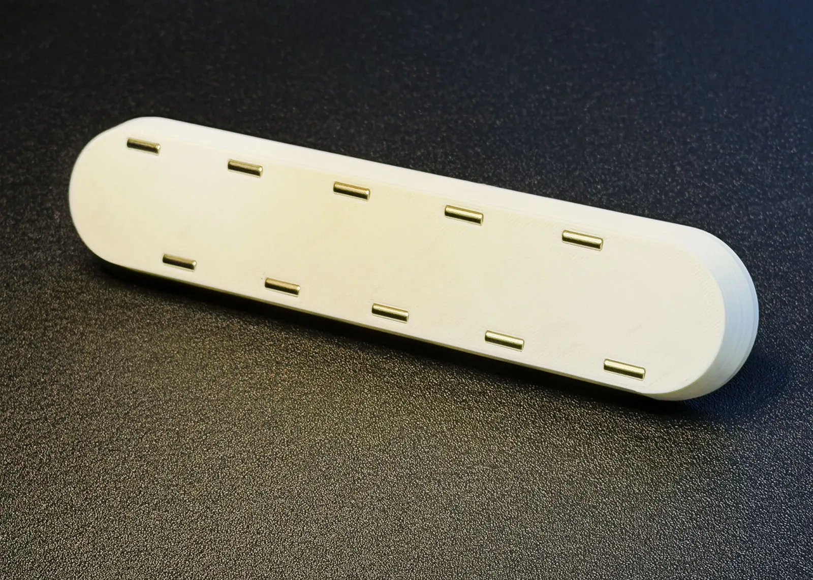 The second white 3D printed bar lying on a dark surface, with 10 magnets lying horizontally in two staggered rows of five, one row near the top edge and one near the bottom