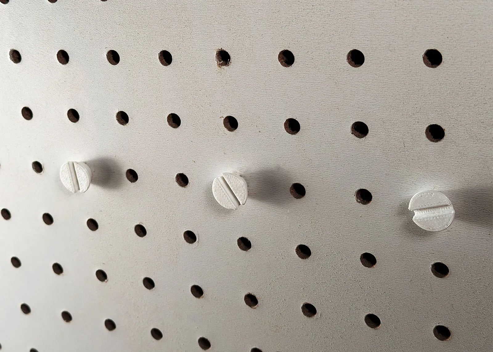 Three white 3D printed slotted screws threaded into a row of pegboard holes, three holes apart, with their heads standing out from the board