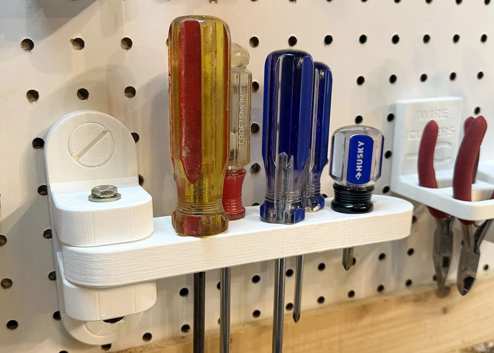 Close-up of the arm folded flat against pegboard, holding five screwdrivers with yellow, clear and blue handles, with the hex bolt hinge pin on top of the mount