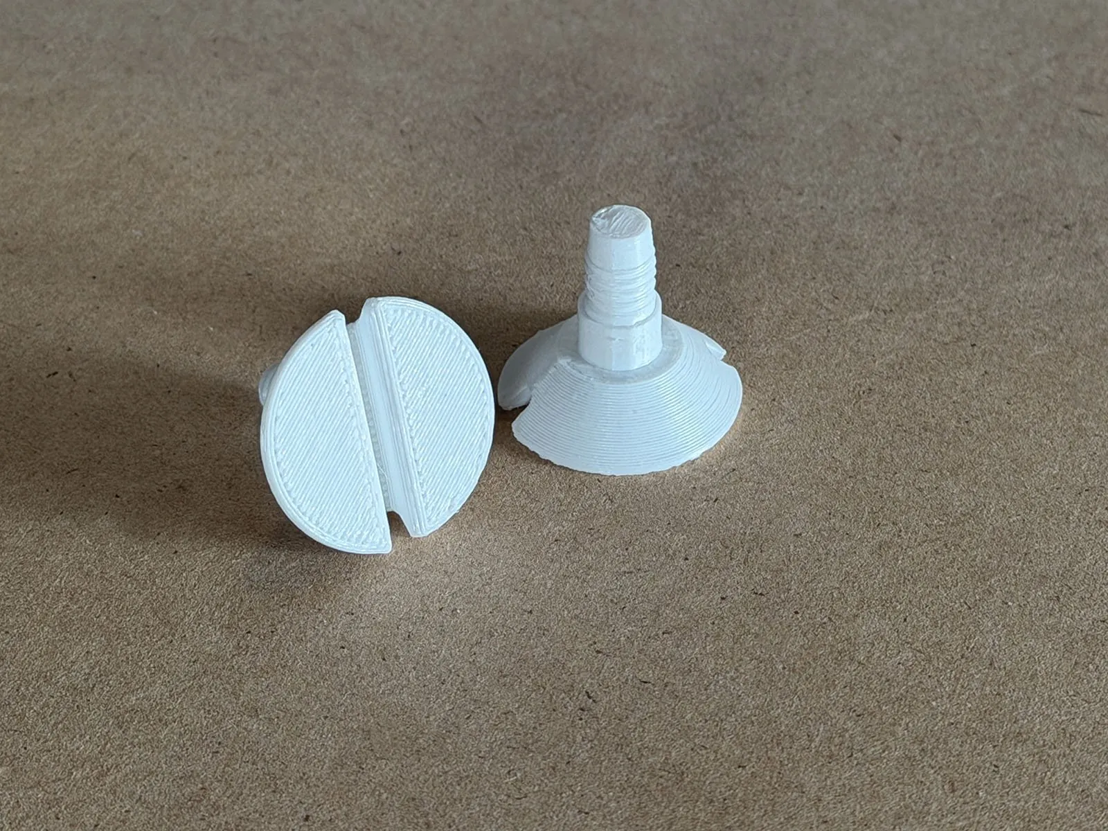 Two white 3D printed 1/4-20 screws on a brown work surface, one showing its slotted countersunk head and the other standing on its head with the thread and tapered tip pointing up