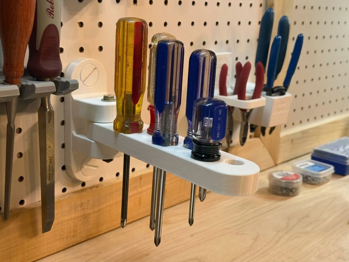 The white printed arm swung out from pegboard above a workbench, holding four screwdrivers with their shafts hanging below it, with pliers holders on the board behind